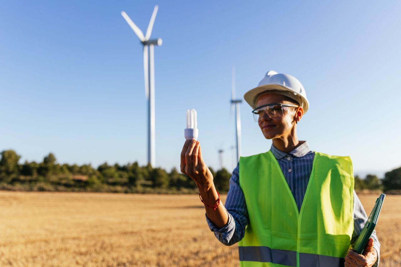 Técnico con casco y chaleco de seguridad junto a aerogeneradores, con una bombilla de bajo consumo en la mano